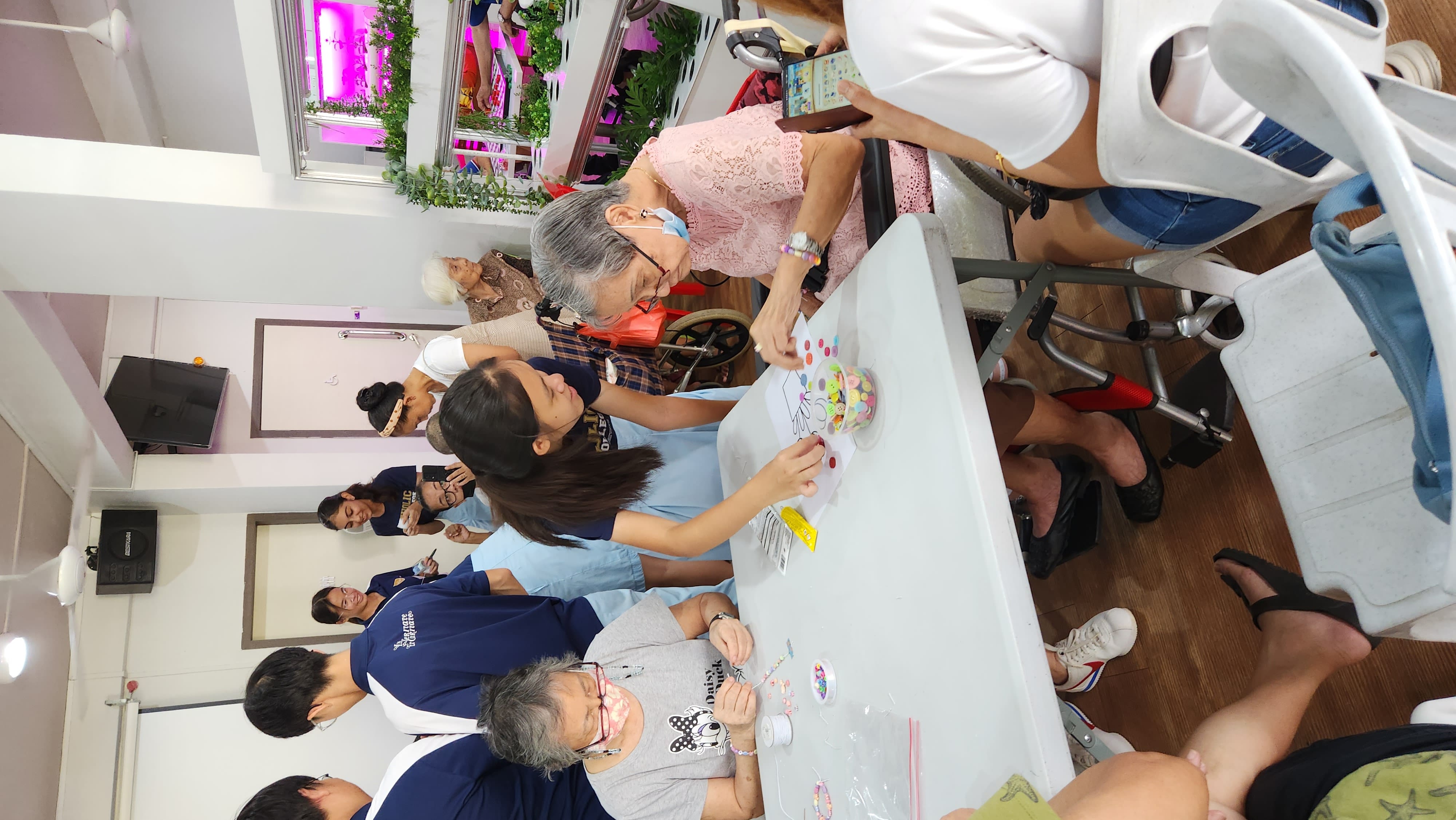 Young volunteers help elderly women with a beading craft activity at a table in a bright indoor community room.